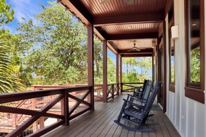 A covered outdoor area with wooden flooring, a rocking chair, and a view of trees and buildings. - Captain Morgan | Steps to Beach | Community Pool | Boardwalk to Beach (WindMark Beach)