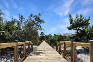 A wooden boardwalk leading through a park with trees on both sides and a clear sky above. - Captain Morgan | Steps to Beach | Community Pool | Boardwalk to Beach (WindMark Beach)