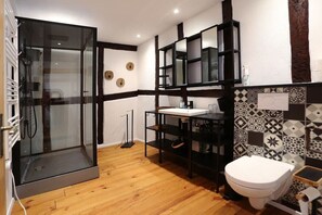 A bathroom with a glass shower enclosure, a white toilet, a sink with a black countertop, and patterned tiles on the walls. - Quartier Blanc de Noir (Reil)