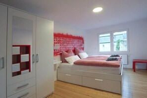 A bedroom with a bed, a wardrobe, a window, and a red bench. - Quartier Blanc de Noir (Reil)