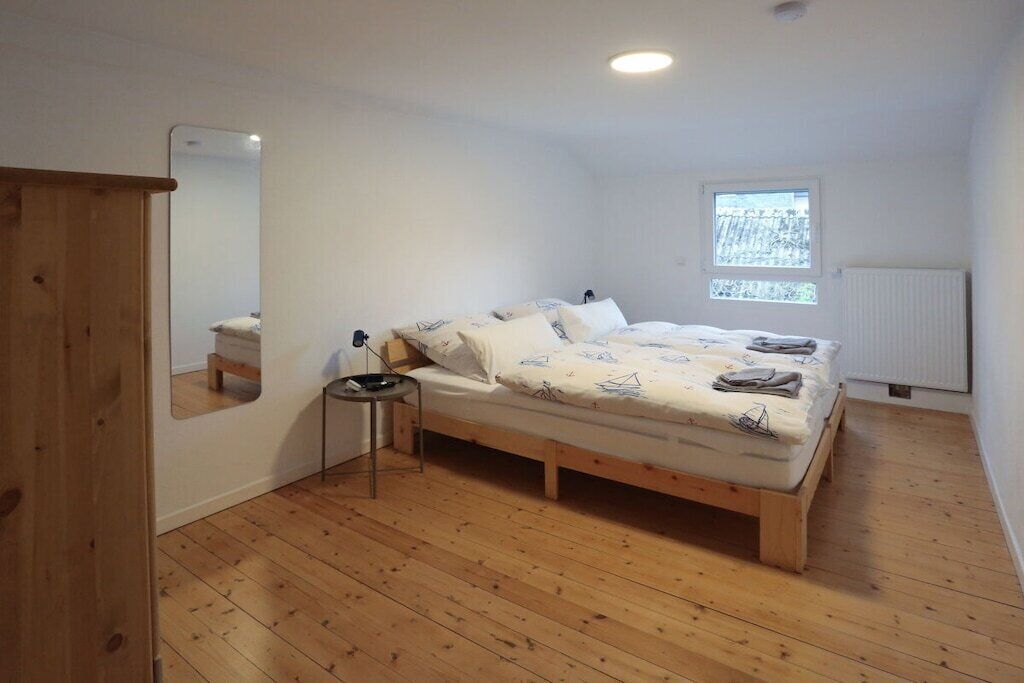 A bedroom with a wooden floor, a bed with a patterned duvet, a small bedside table, a mirror, and a window with a view of trees.