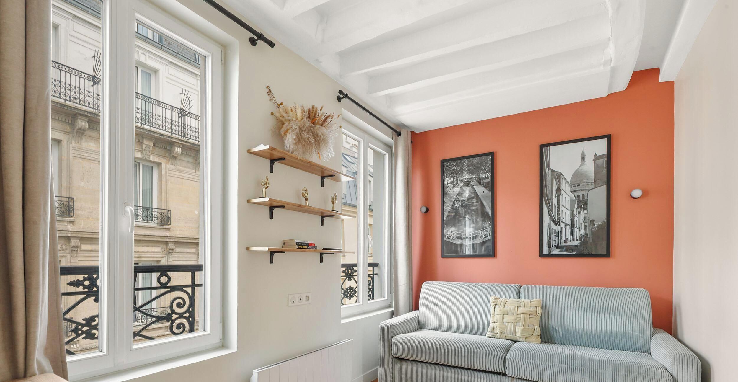 Een kamer met een bank, een radiator, een raam met uitzicht op een gebouw en twee ingelijste foto's aan de muur.