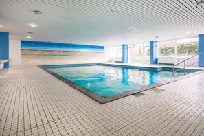 Une piscine intérieure avec un sol carrelé, une vue sur la plage à travers la paroi et un plafond doté d’un éclairage encastré.