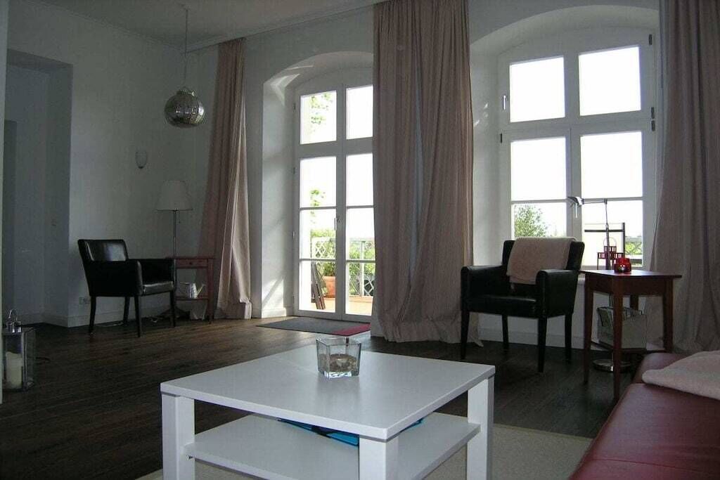 Una sala de estar con una mesa blanca, dos sillas negras, un sofá rojo y ventanas grandes con cortinas.