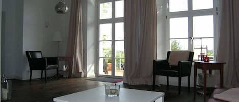 A living room with a white table, two black chairs, a red sofa, and large windows with curtains.