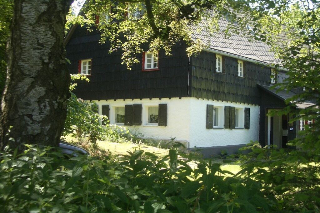 A two-story house with a white exterior and dark shutters, surrounded by greenery.