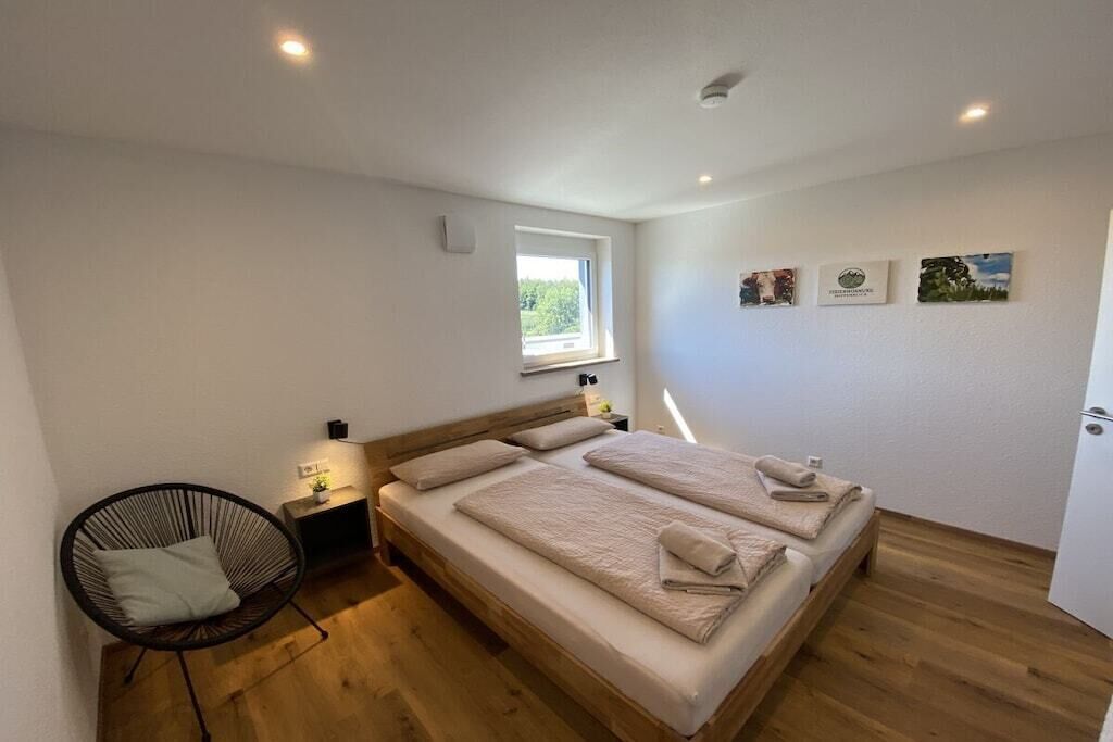 A bedroom with a wooden floor, a bed with beige bedding, a chair, a bedside table with a lamp, and a window.