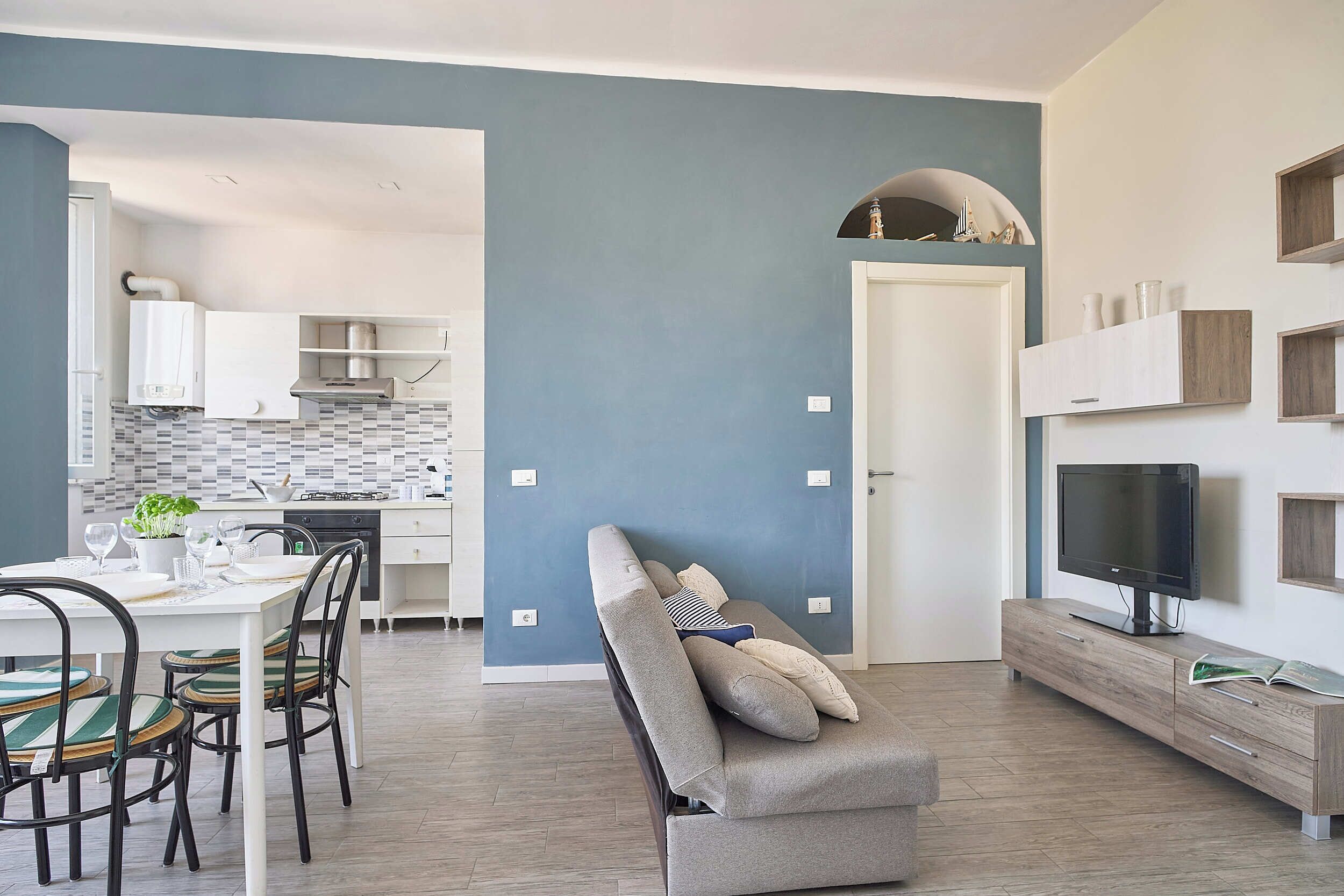 A modern living room with a grey sofa, a dining area with a white table and chairs, a kitchen with white cabinets and a tiled backsplash, and a TV on a wooden stand.
