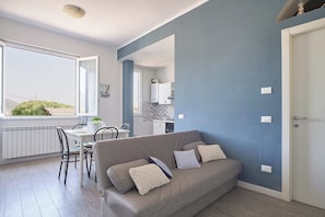 Un salon moderne avec un canapé gris, une table à manger avec des chaises, une cuisine en arrière-plan et une grande fenêtre.