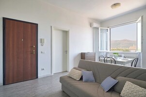 Un soggiorno moderno con una porta in legno, un divano con cuscini decorati, un'area pranzo con tavolo e sedie, e un'ampia finestra che offre vista sul paesaggio.