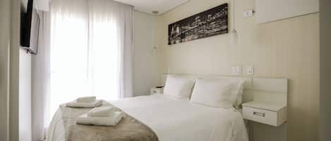 A hotel room with a bed, a bedside table, a wall-mounted air conditioner, a painting, and a window with curtains.