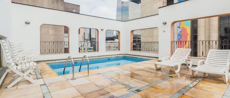 A rooftop pool area with a clear blue pool, white lounge chairs, and a view of the cityscape.