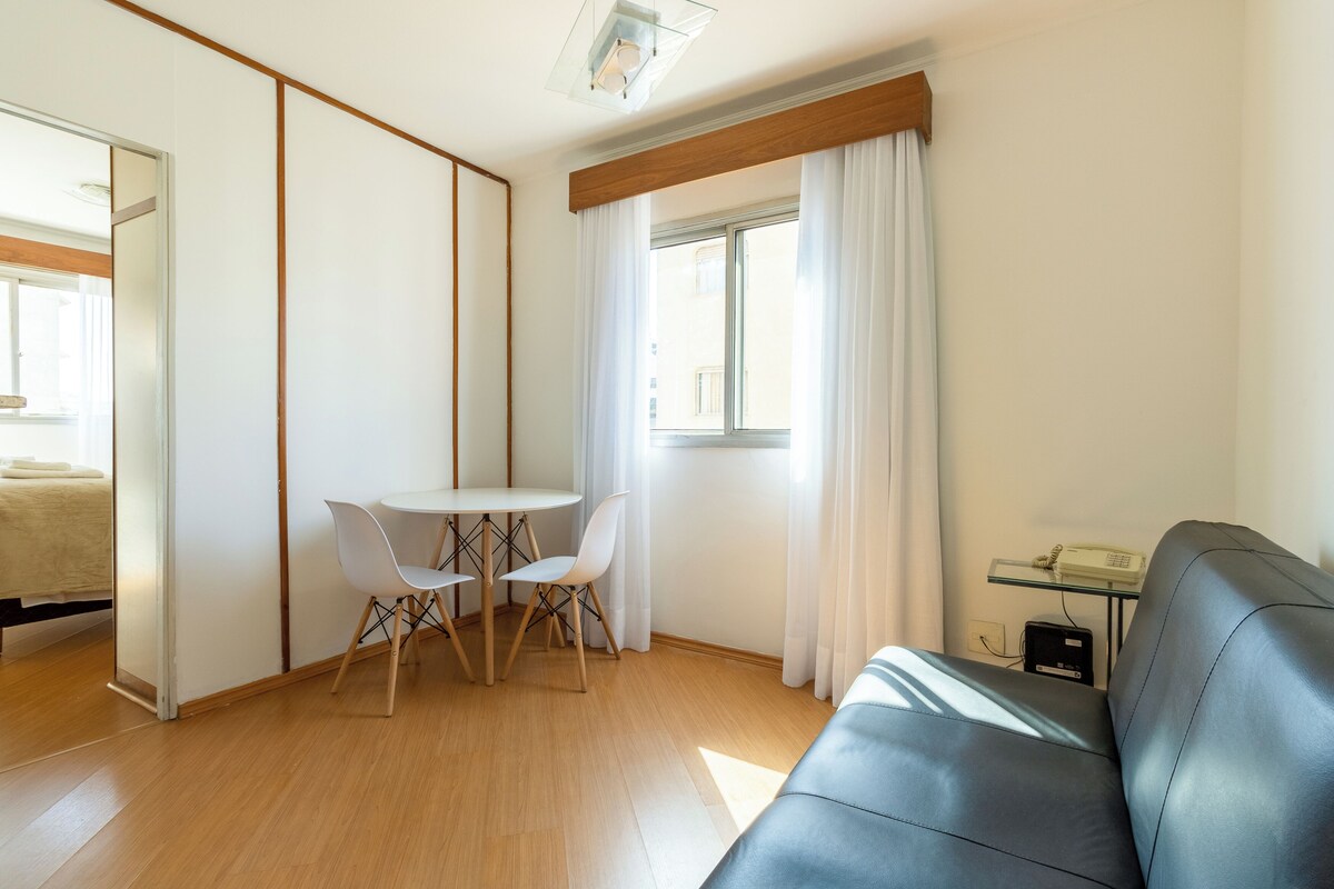 Ein modernes Wohnzimmer mit einer Ledersofa, einem runden Esstisch mit zwei Stühlen, einem Fenster mit weißen Vorhängen und einem sichtbaren Telefon auf einer Glastisch.