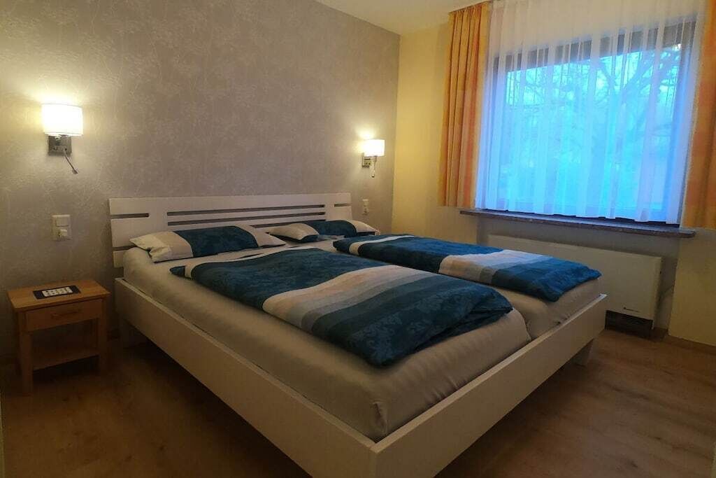 Ein Doppelbett mit Kopfteil, Nachttischlampen, einem Nachttisch und einem fensterbepflanzten Fenster mit Vorhängen.