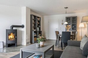 A modern living room with a wood-burning stove, a dining area with a table and chairs, and a built-in bookshelf. - Holiday apartment with 1 bedroom (Fehmarn)