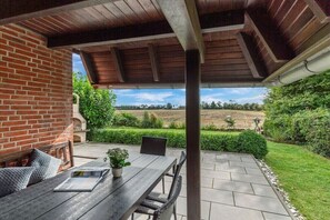 Een terras buiten met een bakstenen muur, een houten tafel met stoelen en uitzicht op een aangelegde tuin.