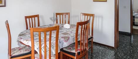 Un comedor con una mesa redonda y seis sillas de madera, una pared blanca con dos cuadros enmarcados, y un piso de baldosas.