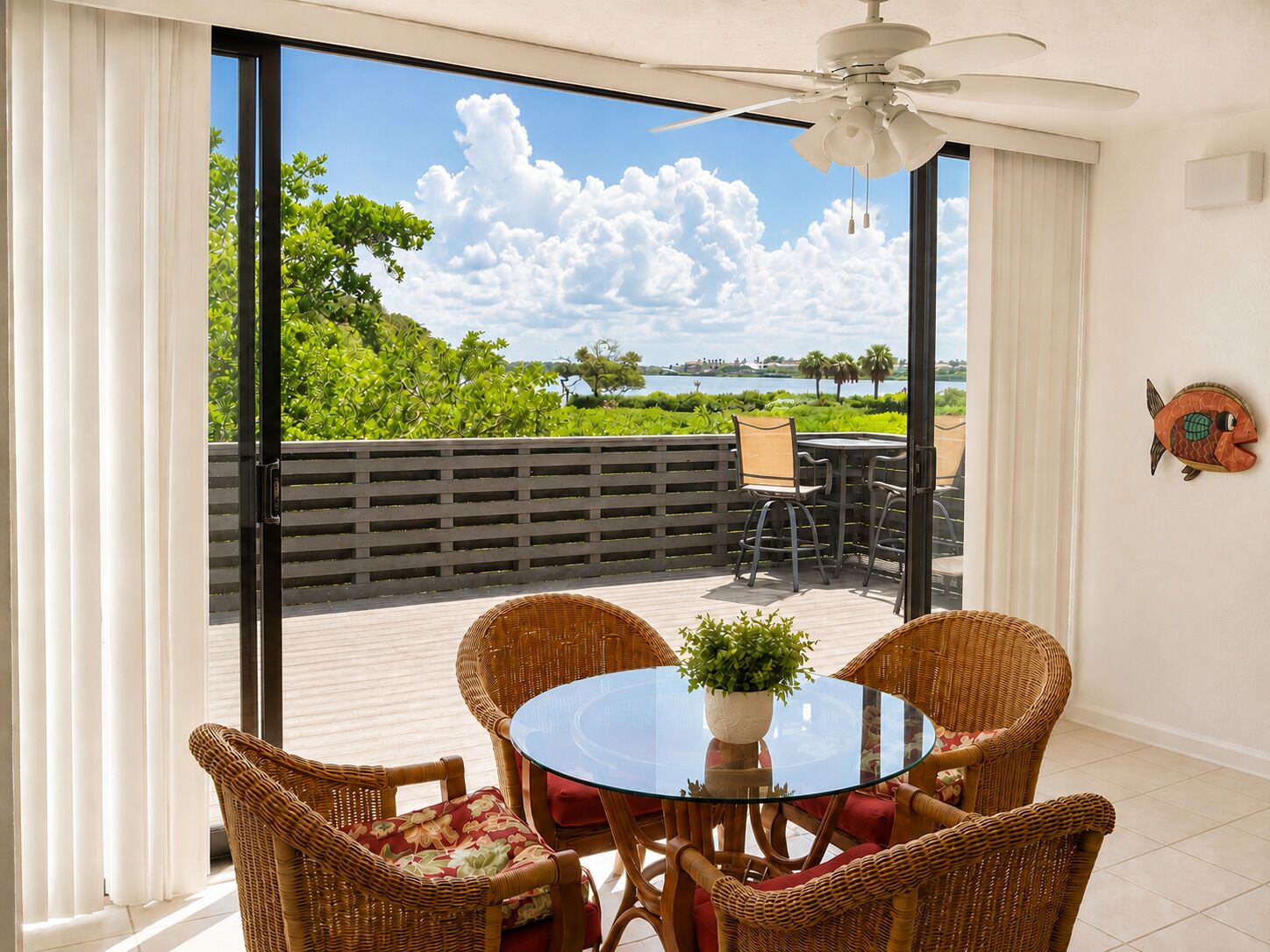 A room with a glass table, wicker chairs, a ceiling fan, and a view of a body of water and greenery.