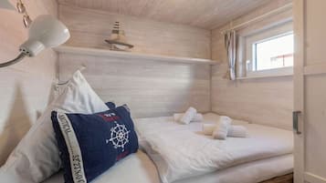 Ein Einzelbett mit weißen Leinen, ein blaues Kopfkissen mit Kompassmuster, eine Wandlampe und ein Fenster mit Vorhängen.
