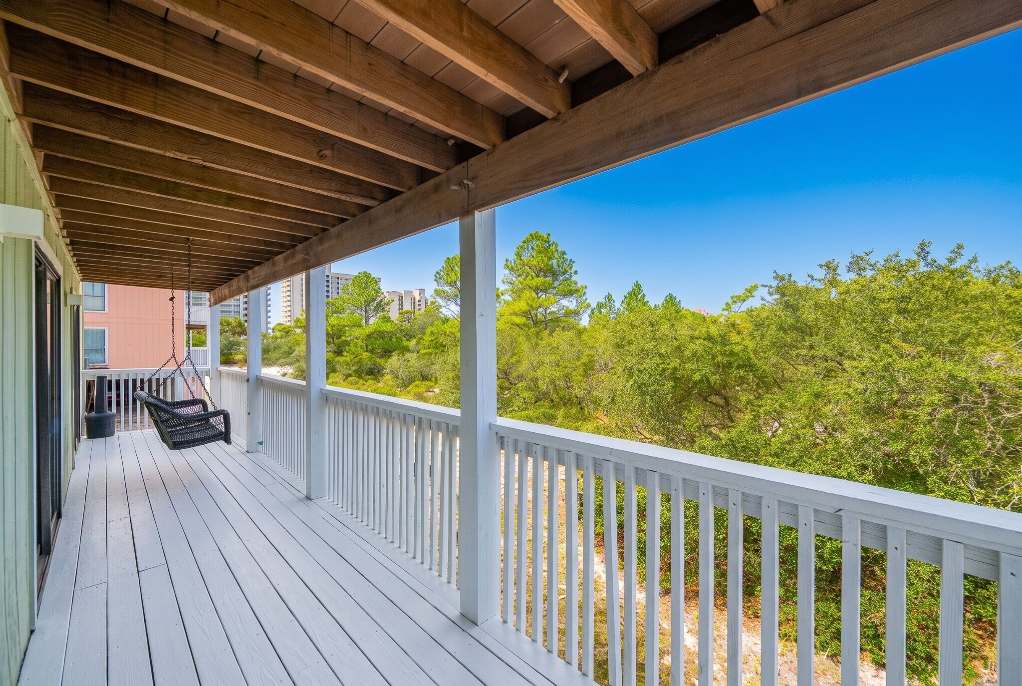 A covered porch with a swing chair, white railings, and a view of green trees and buildings.