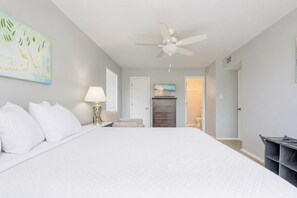 A bedroom with a large bed, a ceiling fan, a nightstand with a lamp, a dresser, and a painting on the wall. - Romar Village 312 2 Bedroom Condo (Orange Beach)