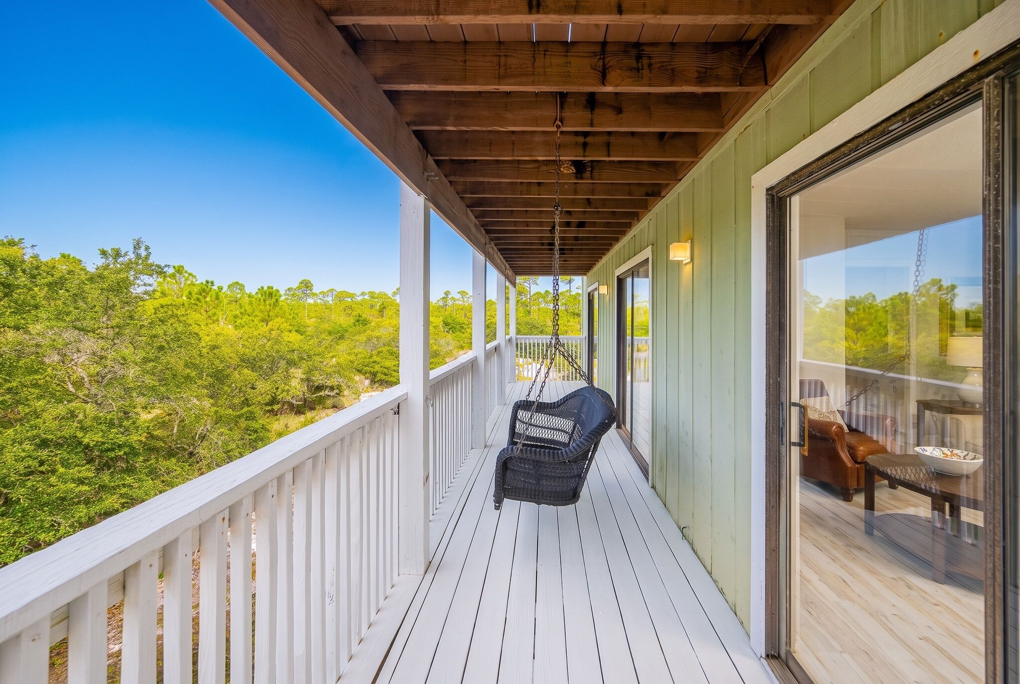 A wooden deck with a swing chair, a glass door leading to an interior space, and a view of green foliage.