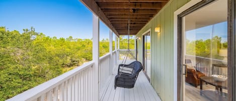 A wooden deck with a swing chair, a glass door leading to an interior space, and a view of green foliage.