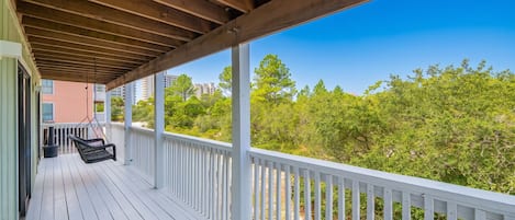 A covered porch with a swing chair, white railings, and a view of green trees and buildings.