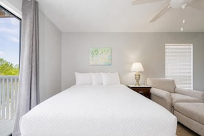 A hotel room with a large bed, a nightstand with a lamp, a chair, a painting on the wall, and a window with blinds. - Romar Village 312 2 Bedroom Condo (Orange Beach)