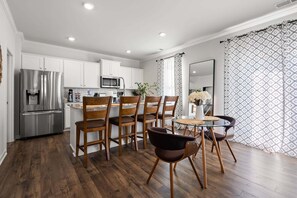 Una cocina moderna con armarios blancos, un refrigerador de acero inoxidable, un microondas y un comedor con sillas de madera y una mesa de vidrio.