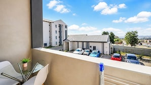 Uma varanda com mesa e cadeiras de vidro, com vista para um estacionamento com vários carros e um prédio ao fundo.