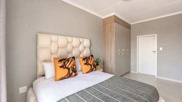 A bedroom with a tufted headboard, a bed with white bedding, two patterned pillows, a grey striped rug, and a wardrobe with a wooden finish.