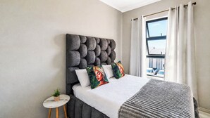 Quarto com cabeceira estofada, cama com roupa de cama branca, mesa lateral redonda pequena com uma planta e vista para um edifício através de uma janela com cortinas.