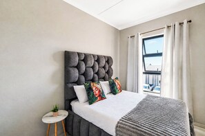 A bedroom with a tufted headboard, a bed with white bedding, a small round side table with a plant, and a view of a building through a window with curtains. - Luxury 2 bedroom, 2 bathroom apartment in Carlswald with swimming pool. (Midrand)