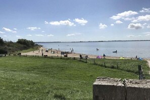 Ein Strand mit einer grasbewachsenen Vordergrundfläche, einem sandigen Bereich mit Menschen und ruhigem Wasser.