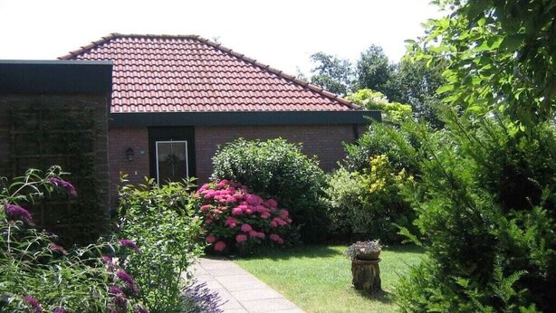 Um edifício residencial com telhado de telhas vermelhas, fachada de tijolos e um jardim frontal com diversas plantas e um caminho.
