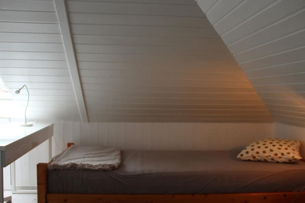 Une petite chambre avec un lit simple, un plafond en pente, une table de chevet et un oreiller.