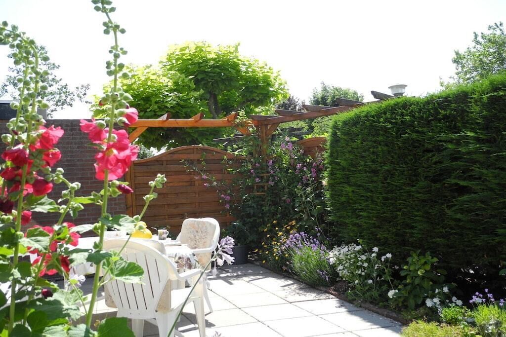 A patio with a wooden pergola, white chairs, and a variety of plants.
