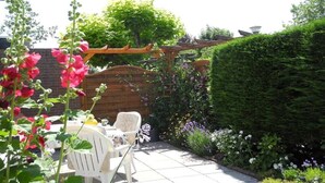 Um pátio com pérgola de madeira, cadeiras brancas e diversas plantas.