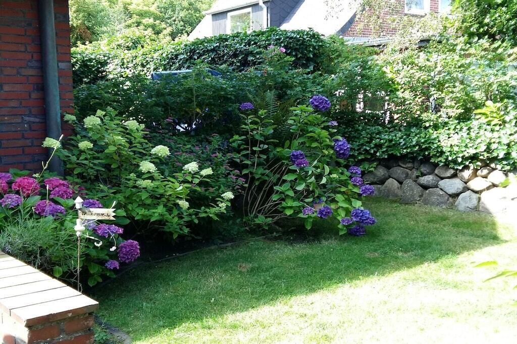 Un jardin avec un mur en briques, une pancarte d’accueil et une variété de plantes, notamment des hortensias.
