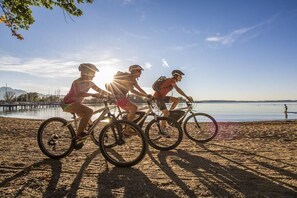 Tre cyklister på en strand med ett vattendrag och en brygga i bakgrunden.
