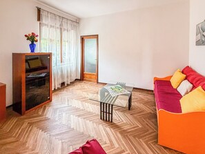 A living room with a herringbone floor, a glass coffee table, a sofa with cushions, a cabinet with a vase, and a door with a window. - A5minutidallagodorta (Gozzano)