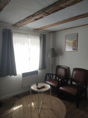Ein Zimmer mit Holzboden, einem fenster mit Vorhängen, einer Lampe, zwei Stühlen, einem kleinen Tisch mit Fernbedienung und einem gerahmten Bild an der Wand.