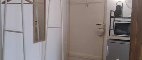 Una habitación con piso de madera, una puerta blanca con un letrero de no fumar, un estante metálico, una silla de madera y un refrigerador pequeño.