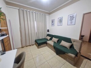 A living room with a sofa, a small table, and a chair. There are curtains on the window, and three framed pictures on the wall.