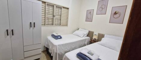 A hotel room with two beds, a wardrobe, a window with blinds, and a wall with framed artwork.