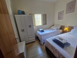 A room with two beds, a wardrobe, a window with blinds, and a wall with a framed picture.