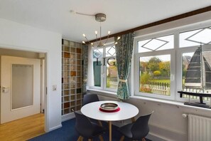 A room with a round table, chairs, a window with curtains, and a view of a white fence and trees. - Holiday apartment Palladium 4 (Langeoog)