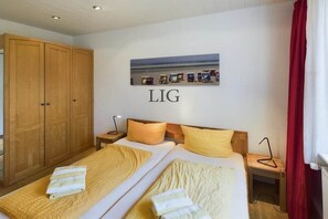 A double bed with two pillows, a wooden wardrobe, a nightstand with a lamp, and a wall-mounted picture. - Holiday apartment Palladium 4 (Langeoog)