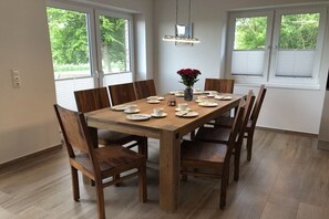 Een eetruimte met een houten tafel voor zes personen, omringd door zes stoelen, met een bloemenvaas op de tafel en een hanglamp erboven.